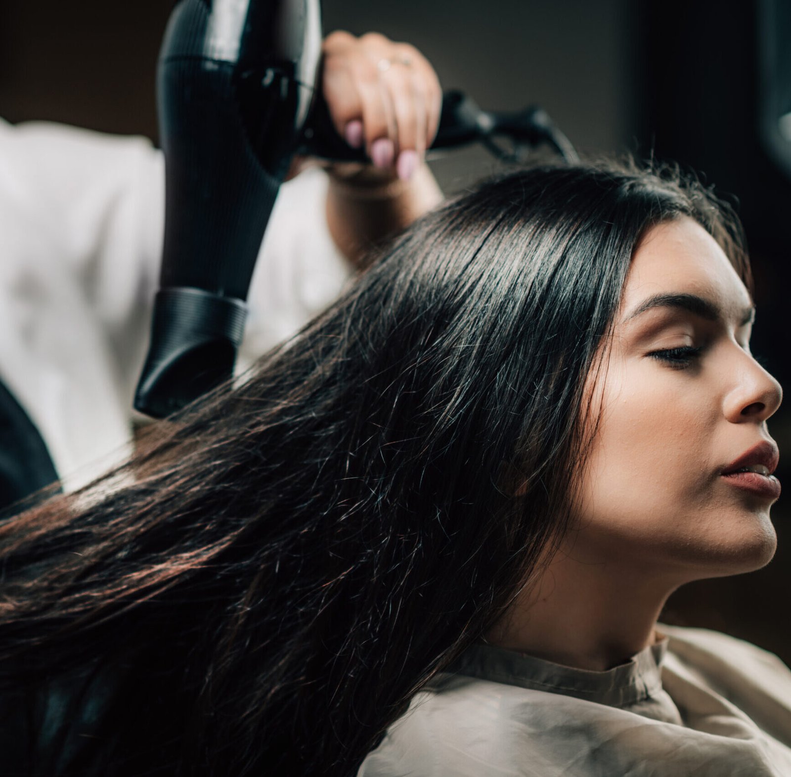 In a salon, a hairdresser blow-dries a woman's long hair, showcasing a professional styling session.