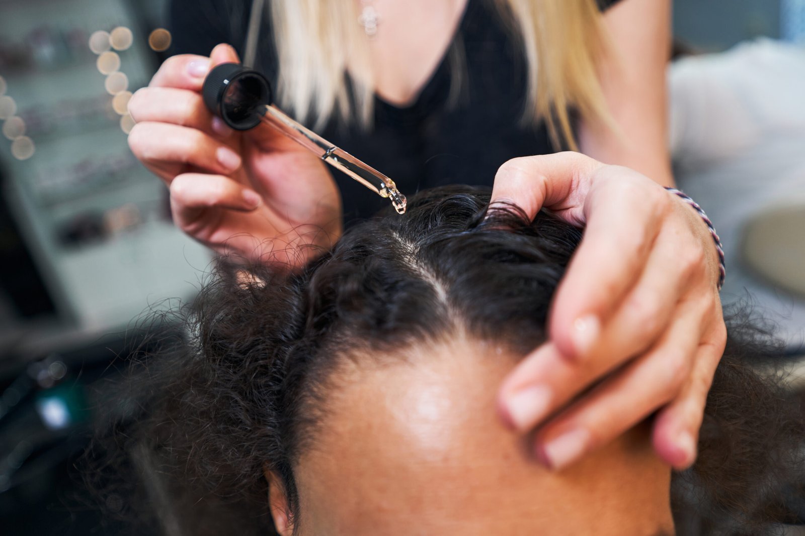 A woman receives pampering in a salon, with a hairdresser applying serum to her hair for a radiant, polished finish.