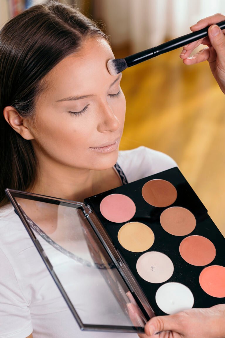 A person is having makeup applied with a brush to their forehead with an open makeup palette showcasing different shades.