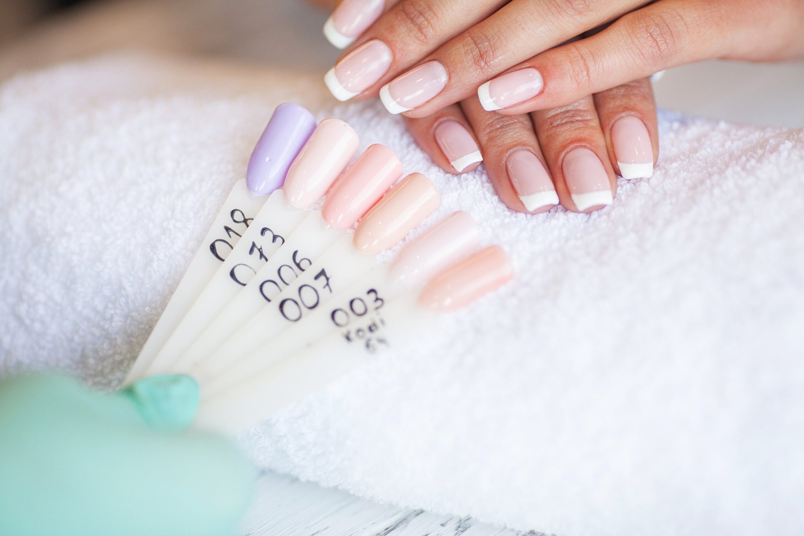 Hands with classic French tip nails resting on a towel beside a nail color palette showcasing pastel gel polish options in a nail salon.