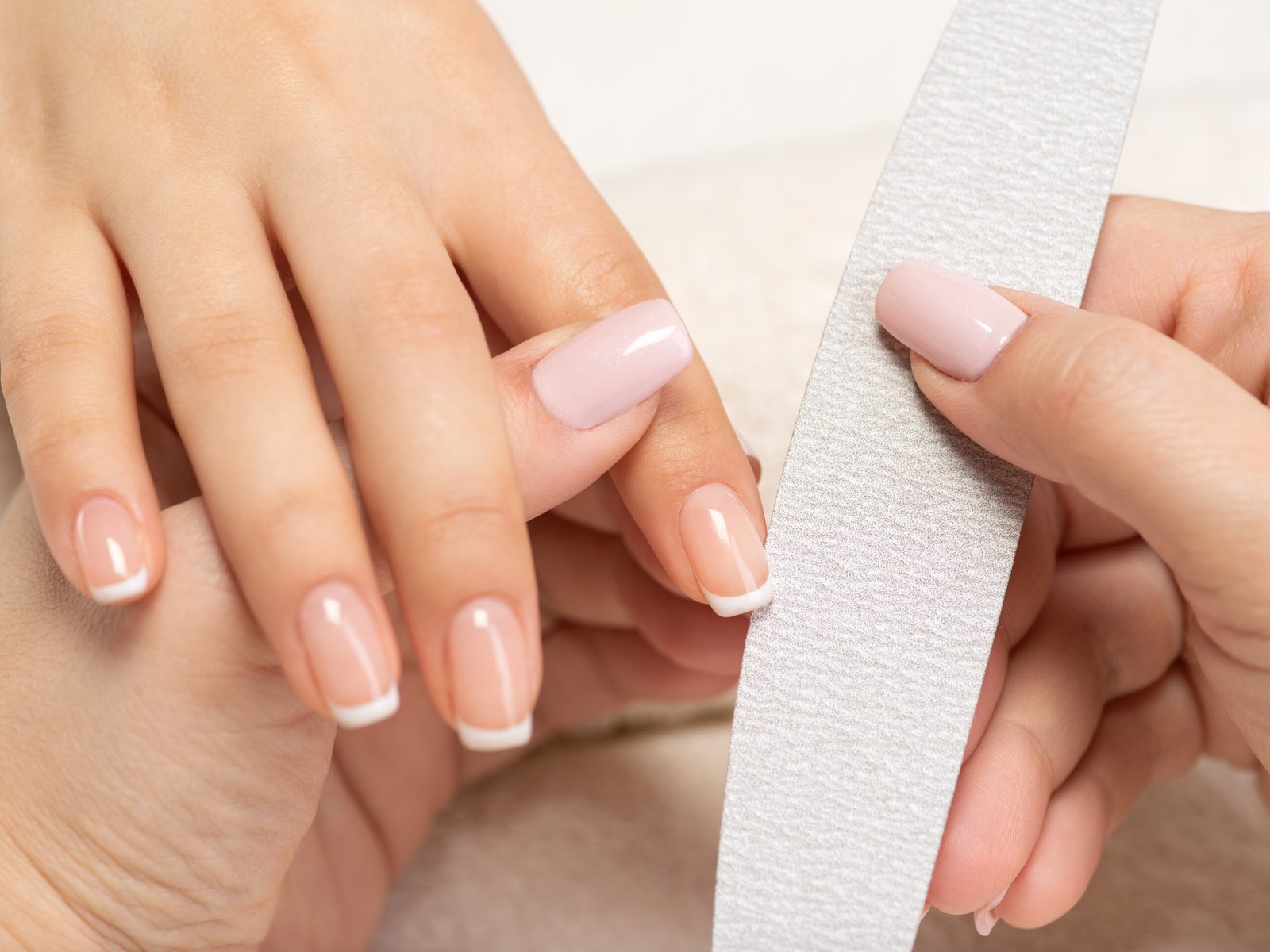 Close-up of a professional French manicure with natural pink base and white tips, nail technician filing nails in a salon setting.