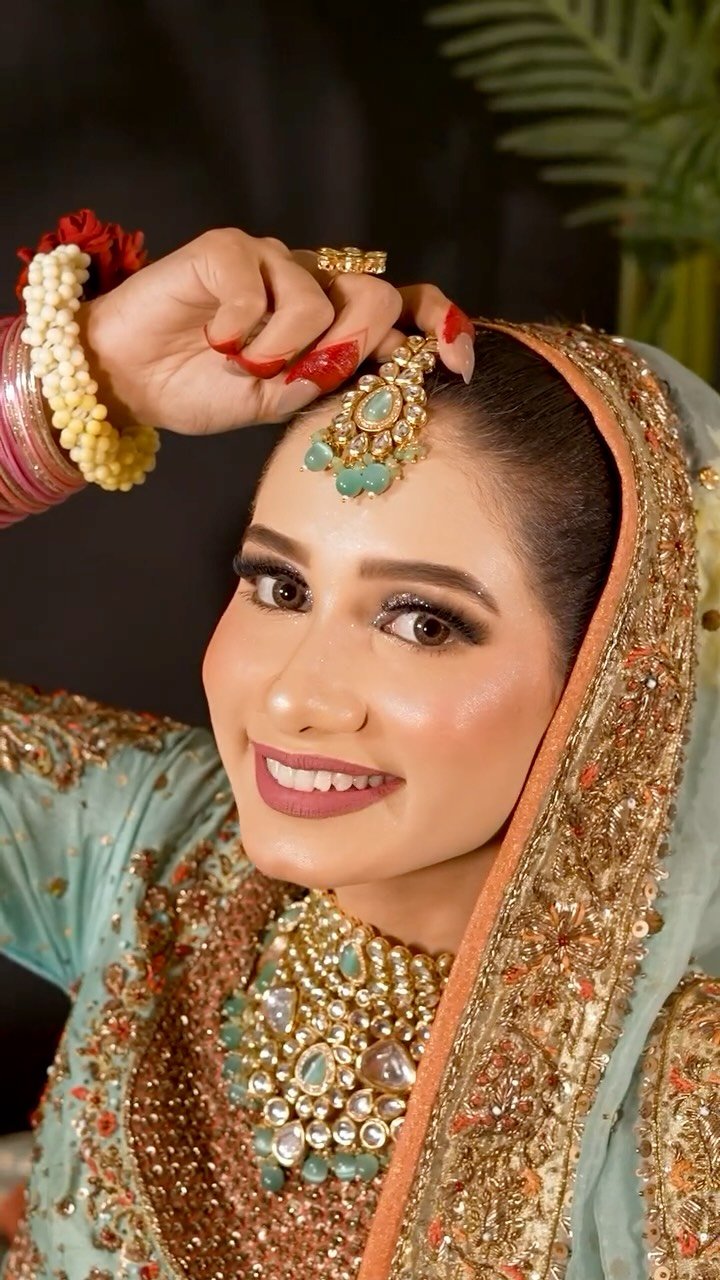 A bride with warm-toned makeup and traditional South Asian jewelry, featuring a vibrant floral wrist accessory.