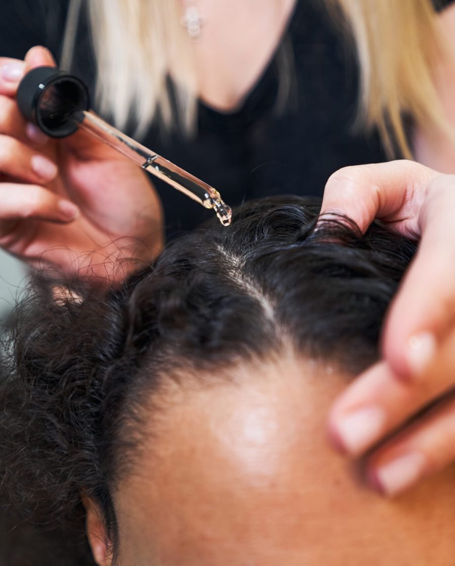A woman receives pampering in a salon, with a hairdresser applying serum to her hair for a radiant, polished finish.