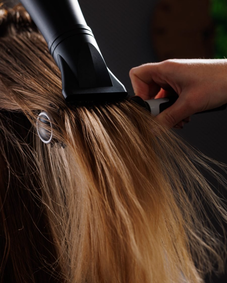 A woman using a blow dryer to style her hair, holding the dryer in one hand with a focused expression.